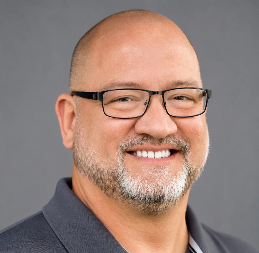 Smiling man with glasses, a short beard, and a bald head, wearing a dark collared shirt, against a plain gray background.
